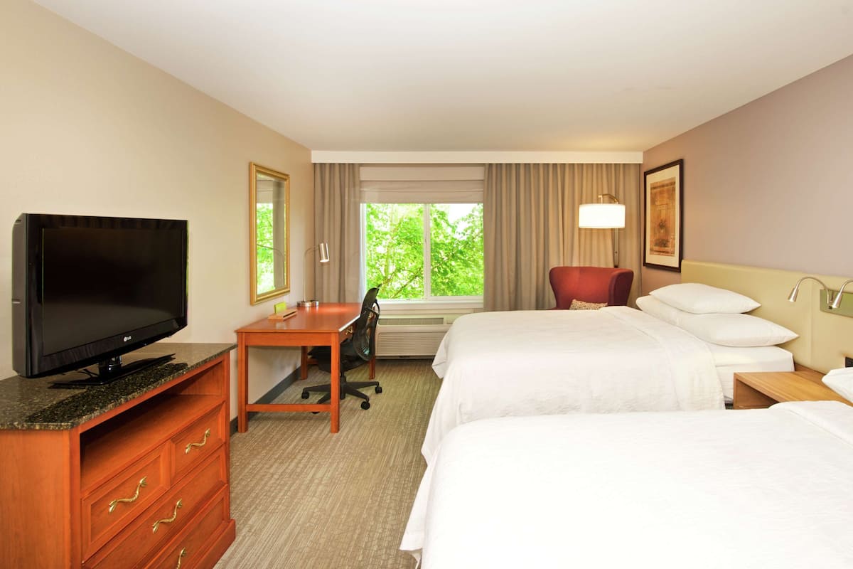 A hotel room with two beds, a desk with a TV, a chair, and a window with curtains.