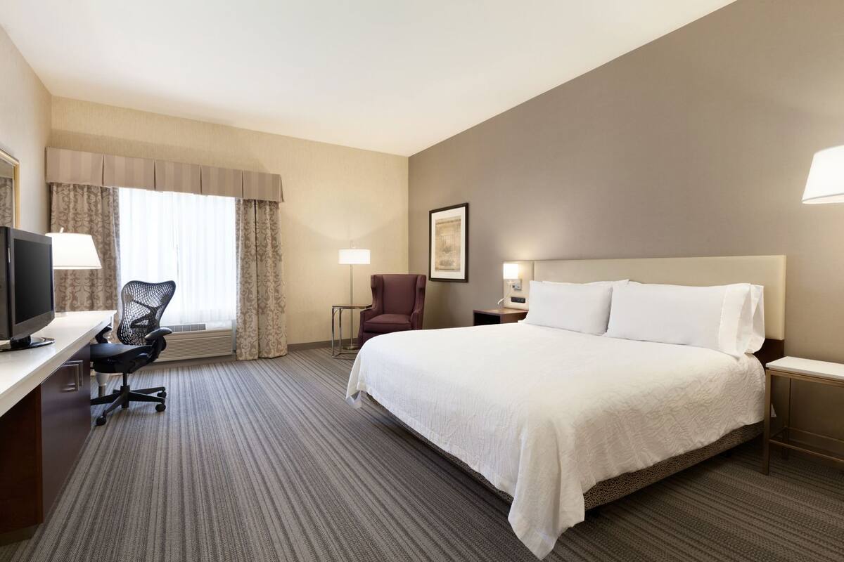 A hotel room with a large bed, a desk with a computer, a chair, a lamp, and a framed picture on the wall.