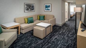 Habitación de hotel con un sofá beige tipo seccional, una mesita, un escritorio y una cama al fondo.