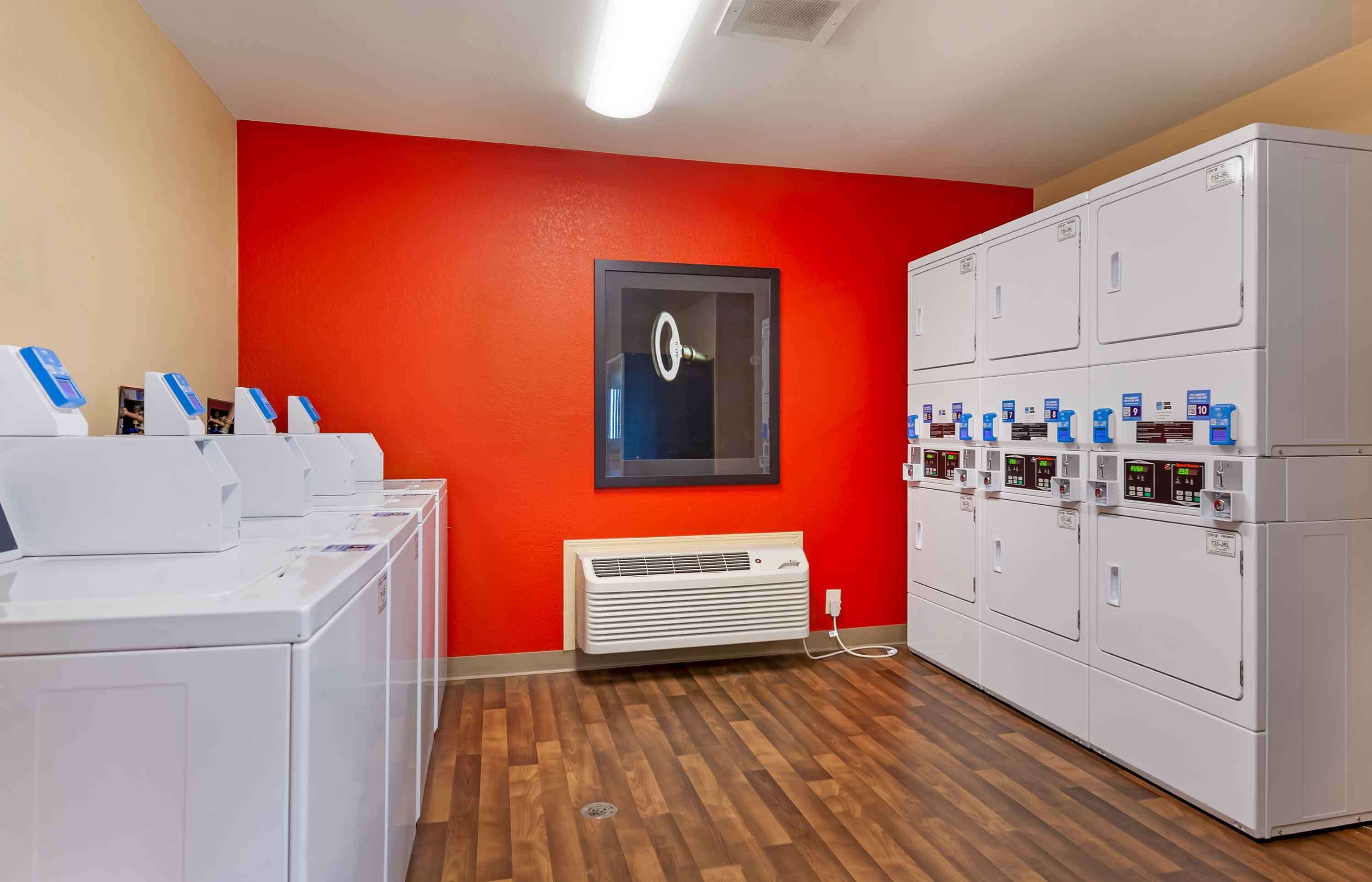 Une buanderie équipée de plusieurs machines à laver et de plusieurs sèche-linge, avec un mur rouge et un tableau encadré.