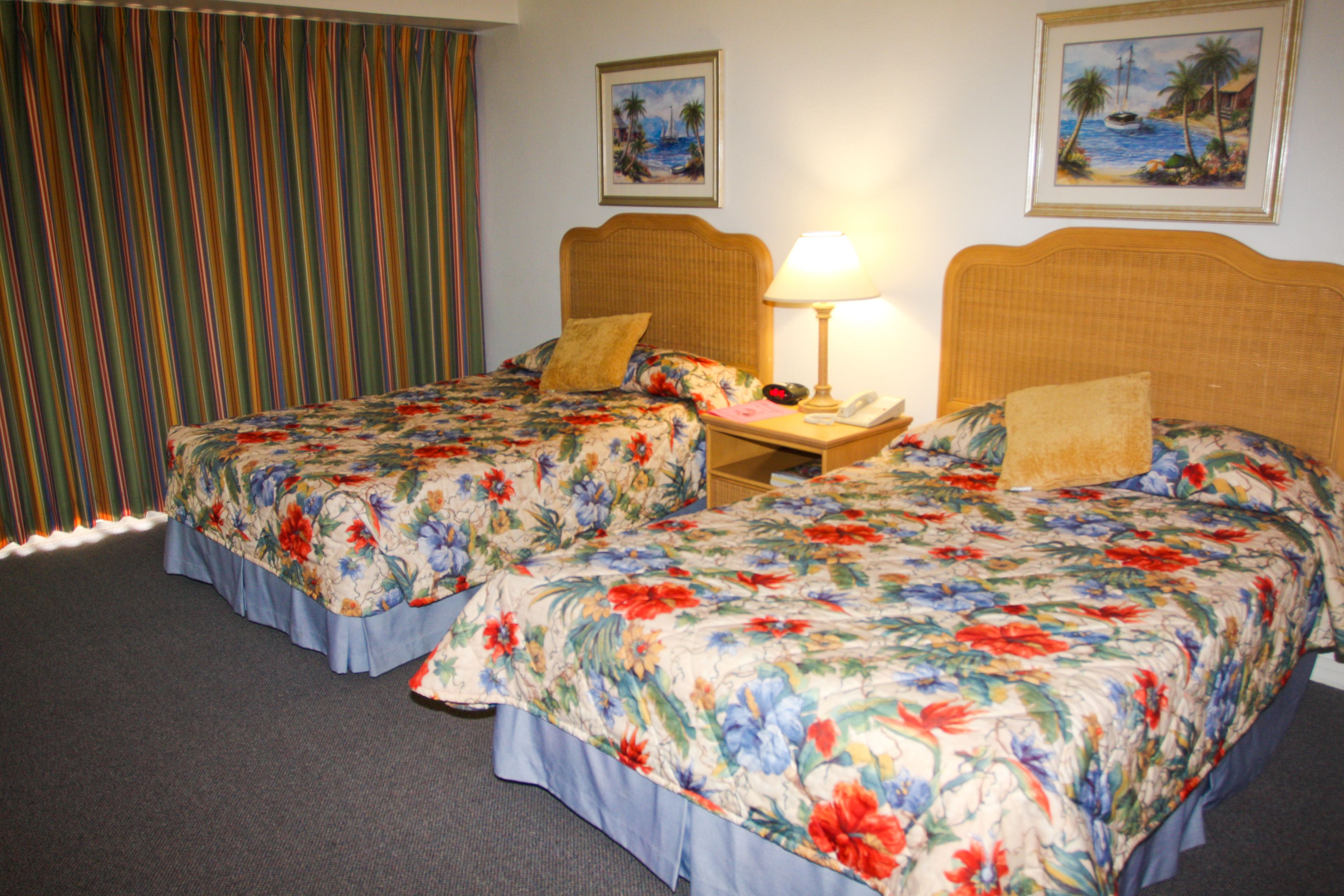 Quarto de hotel com duas camas, cada uma com roupa de cama floral e cabeceira de madeira. Há uma mesa de cabeceira com uma luminária e um telefone entre as camas. O quarto tem cortinas listradas e duas imagens emolduradas na parede.