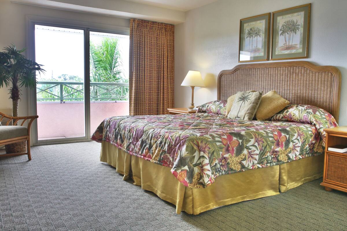 Quarto de hotel com uma cama grande, varanda com vista, cadeira de vime e uma luminária.