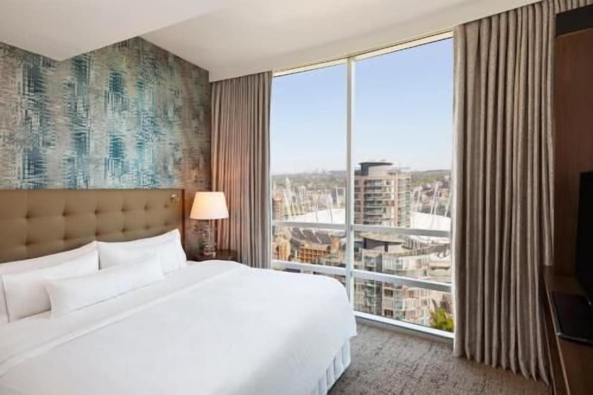 A hotel room with a large bed, a view of the city through floor-to-ceiling windows, and a modern headboard.