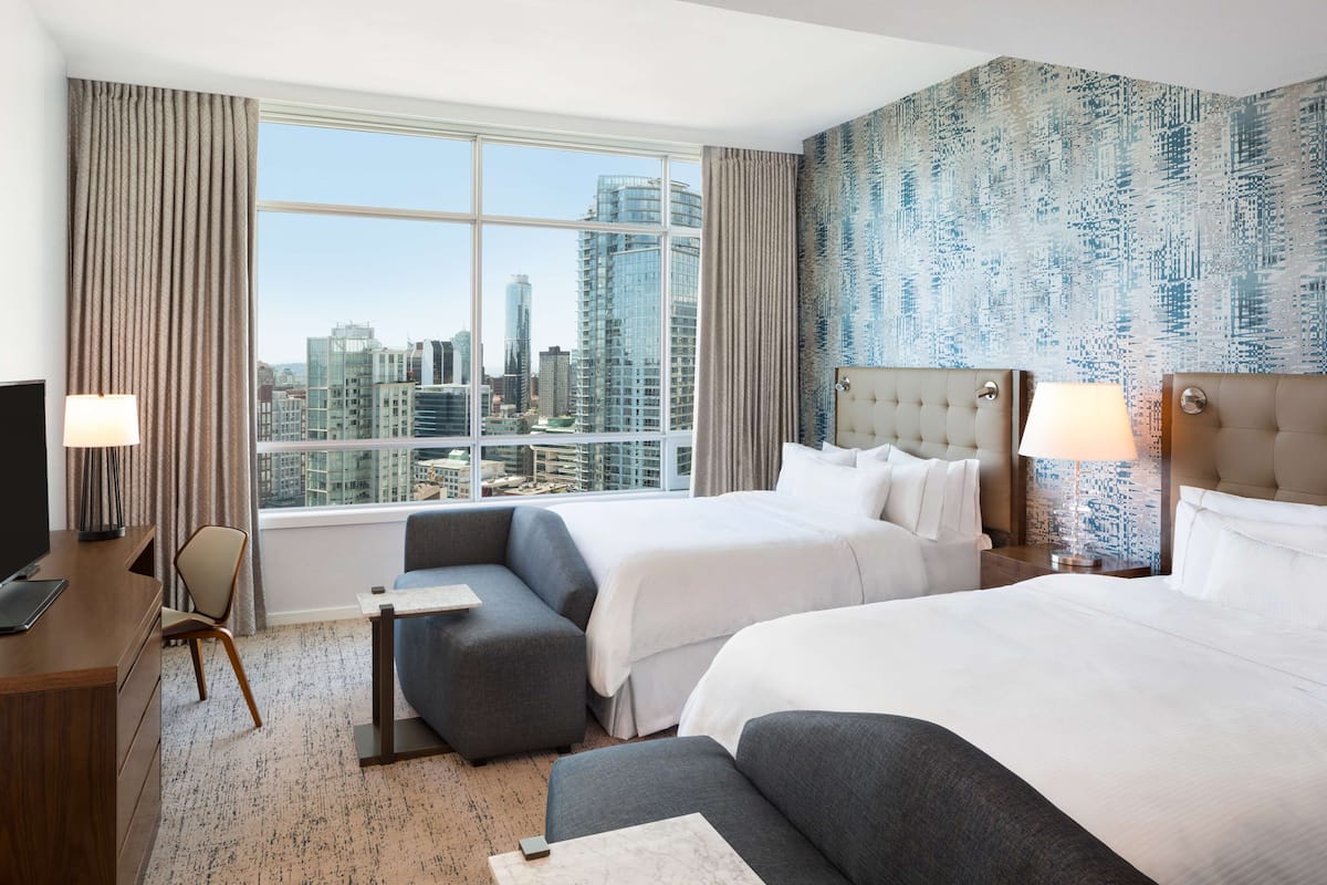A hotel room with two beds, a sofa, a desk, and a large window offering a city view.