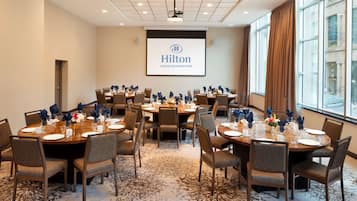 Kokoushuone, jossa on pyöreitä pöytiä, tuoleja ja suuri näyttö, jossa näkyy Hiltonin logo.
