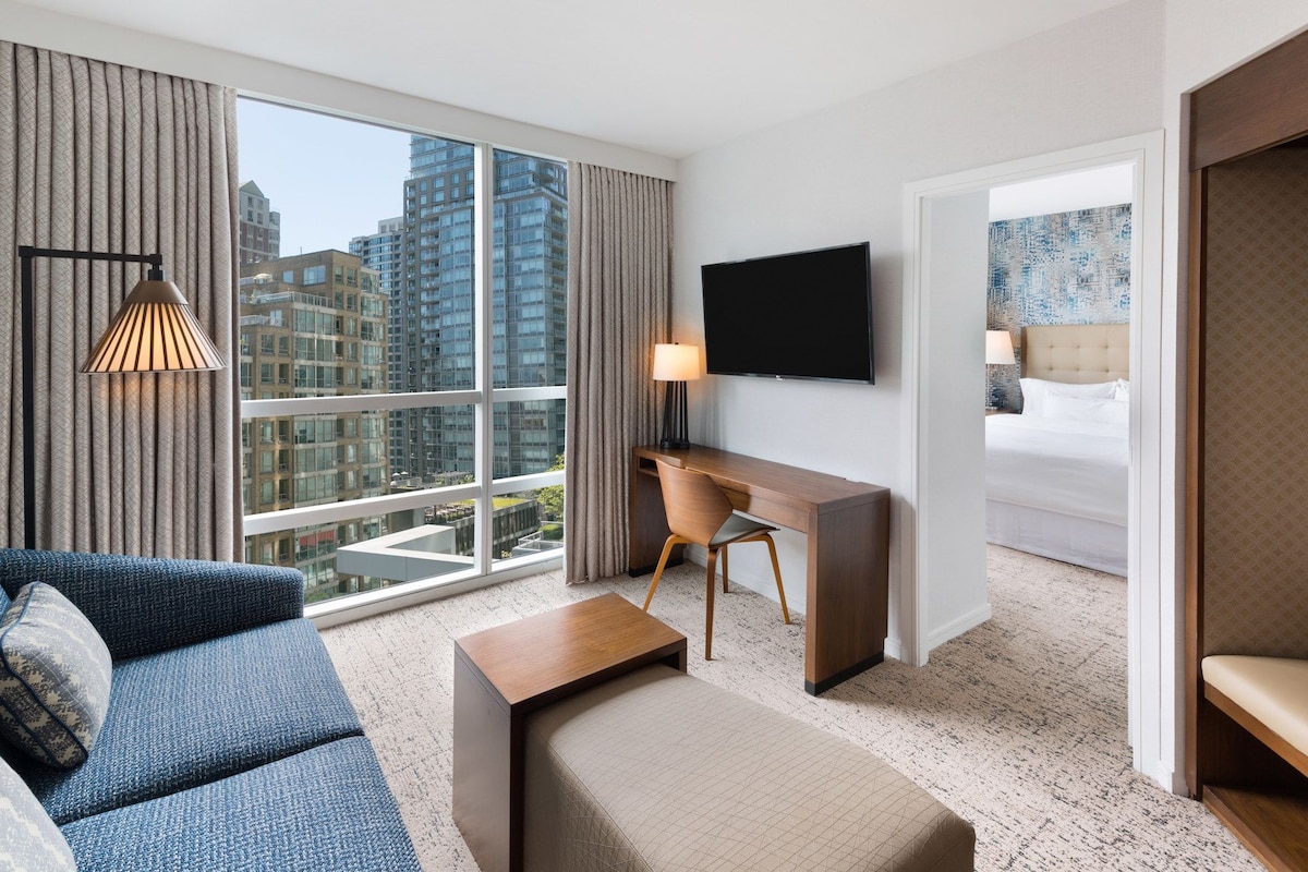 A hotel room with a large window, a desk, a bed, and a sofa.