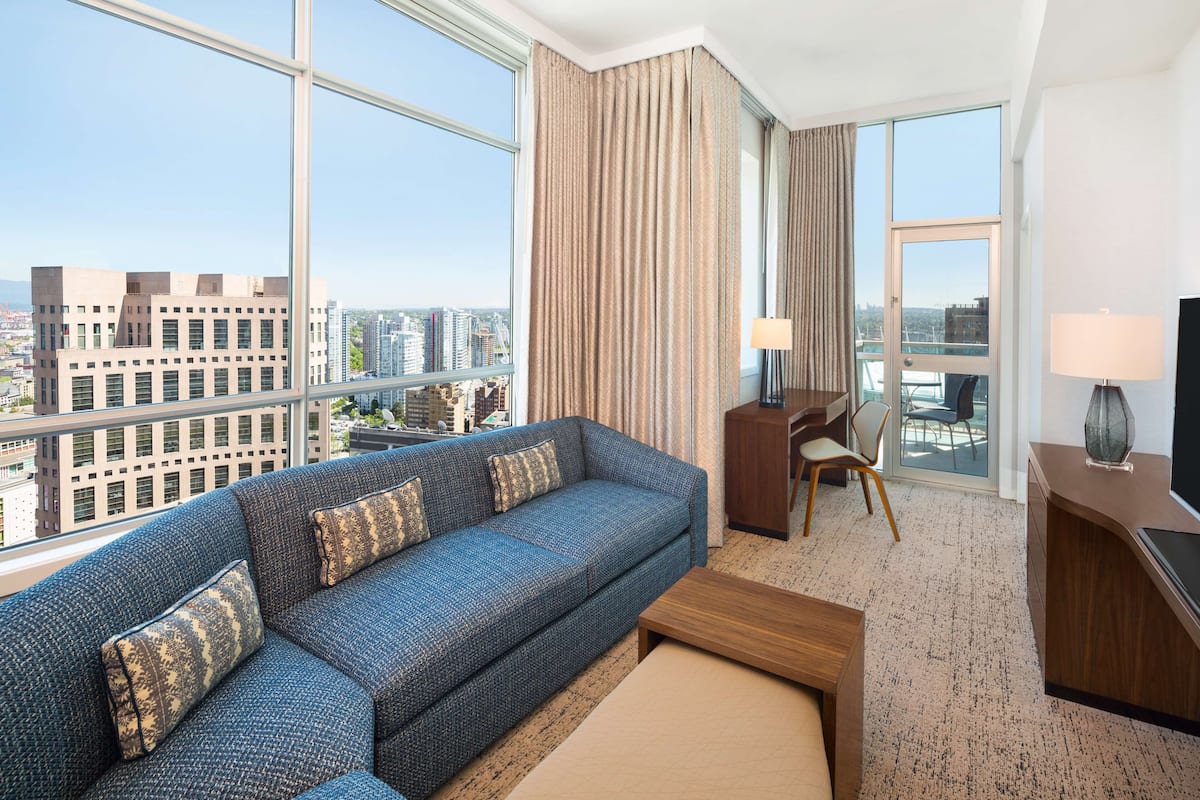 A modern hotel room with a large window offering a city view, a blue sofa with patterned cushions, a wooden coffee table, and a TV on a wooden stand.