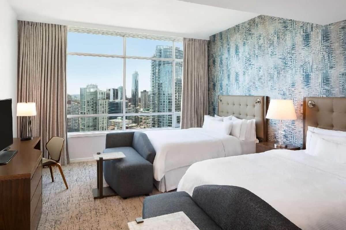 A hotel room with two beds, a sofa, a desk, and a large window with a city view.