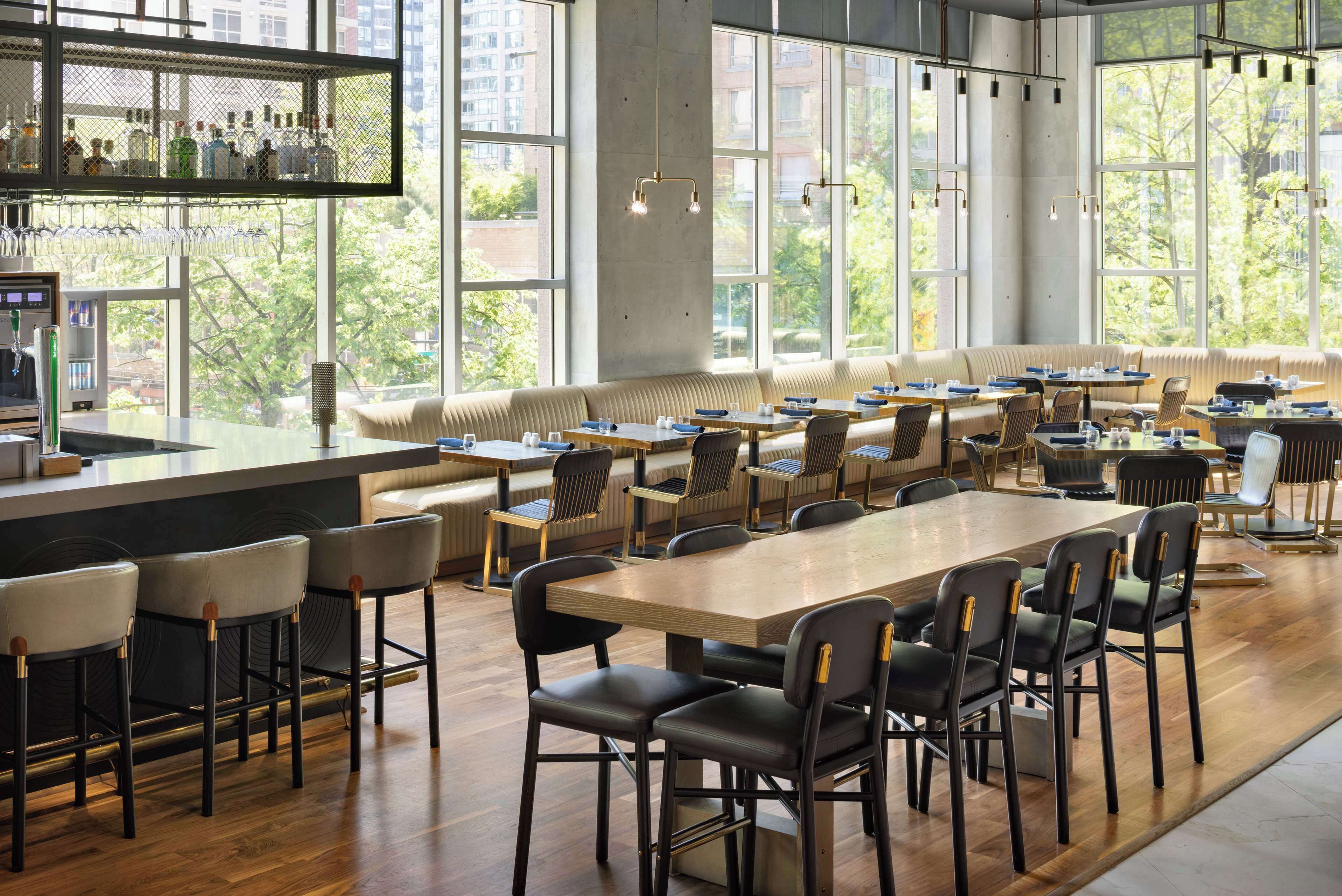 A spacious cafe with a long wooden table, multiple seating areas, and large windows offering a view of the city.