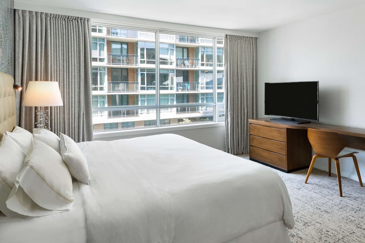 A hotel room with a large bed, a desk, a television, and a window with a view of a building.