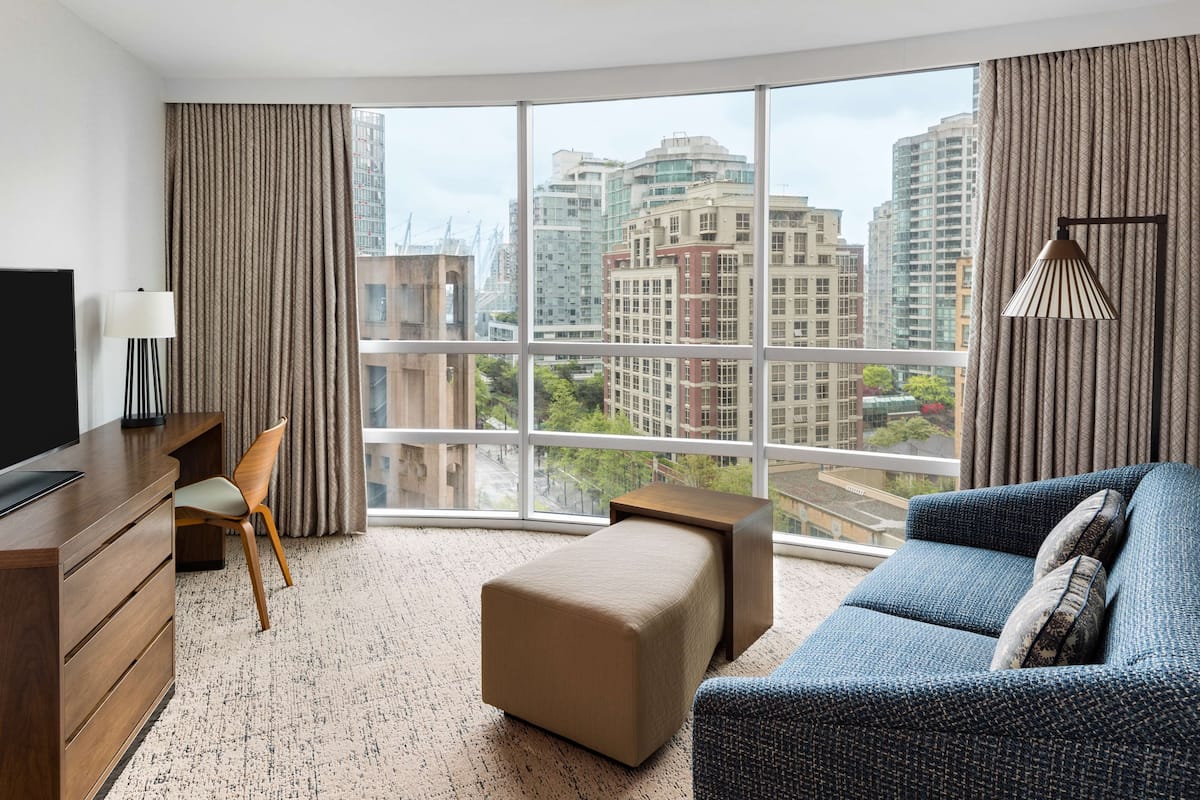 A modern hotel room with a large window, a sofa, a desk, and a TV.