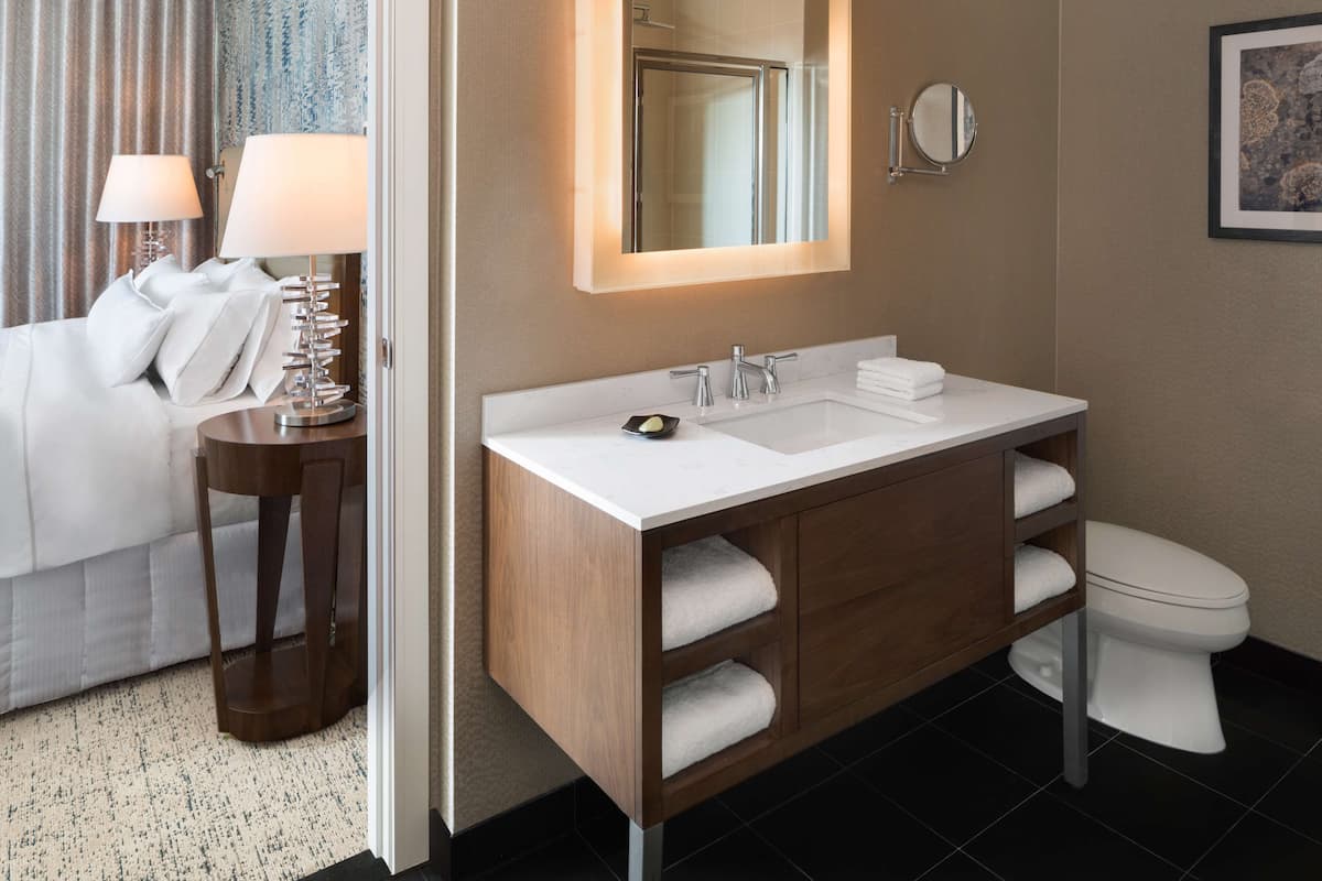A modern bathroom with a white sink, a mirror, and a toilet. There is a towel rack with towels, a small table with a lamp, and a bed with white bedding in the adjacent room.