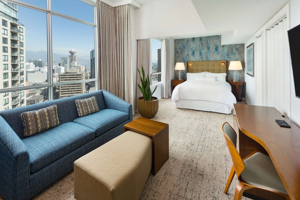 A hotel room with a large window, a bed, a sofa, a desk, and a chair.