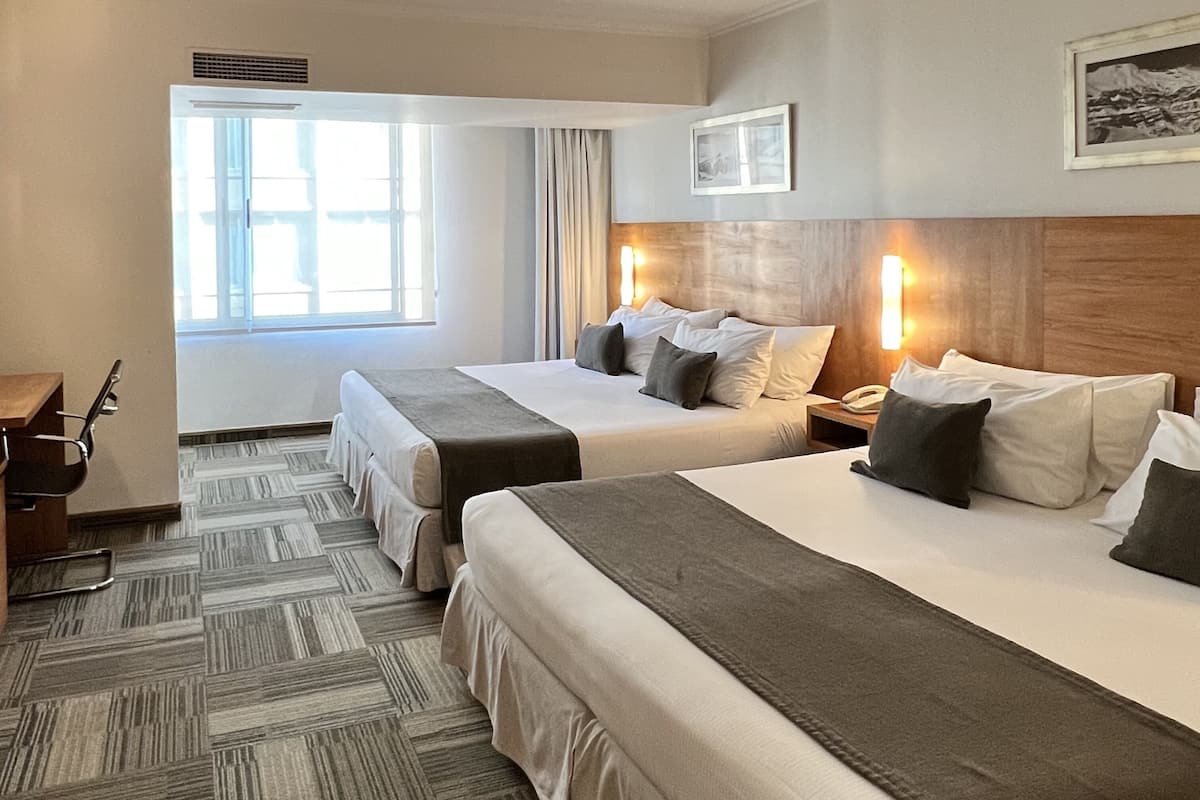 A hotel room with two beds, a desk, and a chair. There is a window with curtains, a framed picture on the wall, and a telephone on the nightstand.
