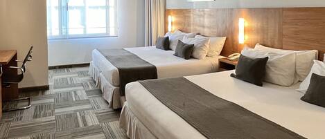 Habitación de hotel con dos camas, un escritorio y una silla. Hay una ventana con cortinas, un cuadro en la pared y un teléfono en la mesita de noche.