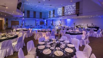 Un salón de banquetes con mesas redondas dispuestas para cenar, sillas blancas y una zona para escenario.