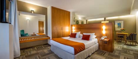 Habitación de hotel con una cama grande, un armario de madera, una mesita de noche con una lámpara, una pequeña mesa con un jarrón y una silla.