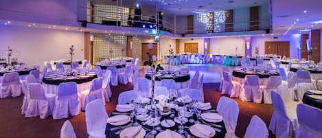 A banquet hall with round tables set for dining, white chairs, and a stage with blue lighting.