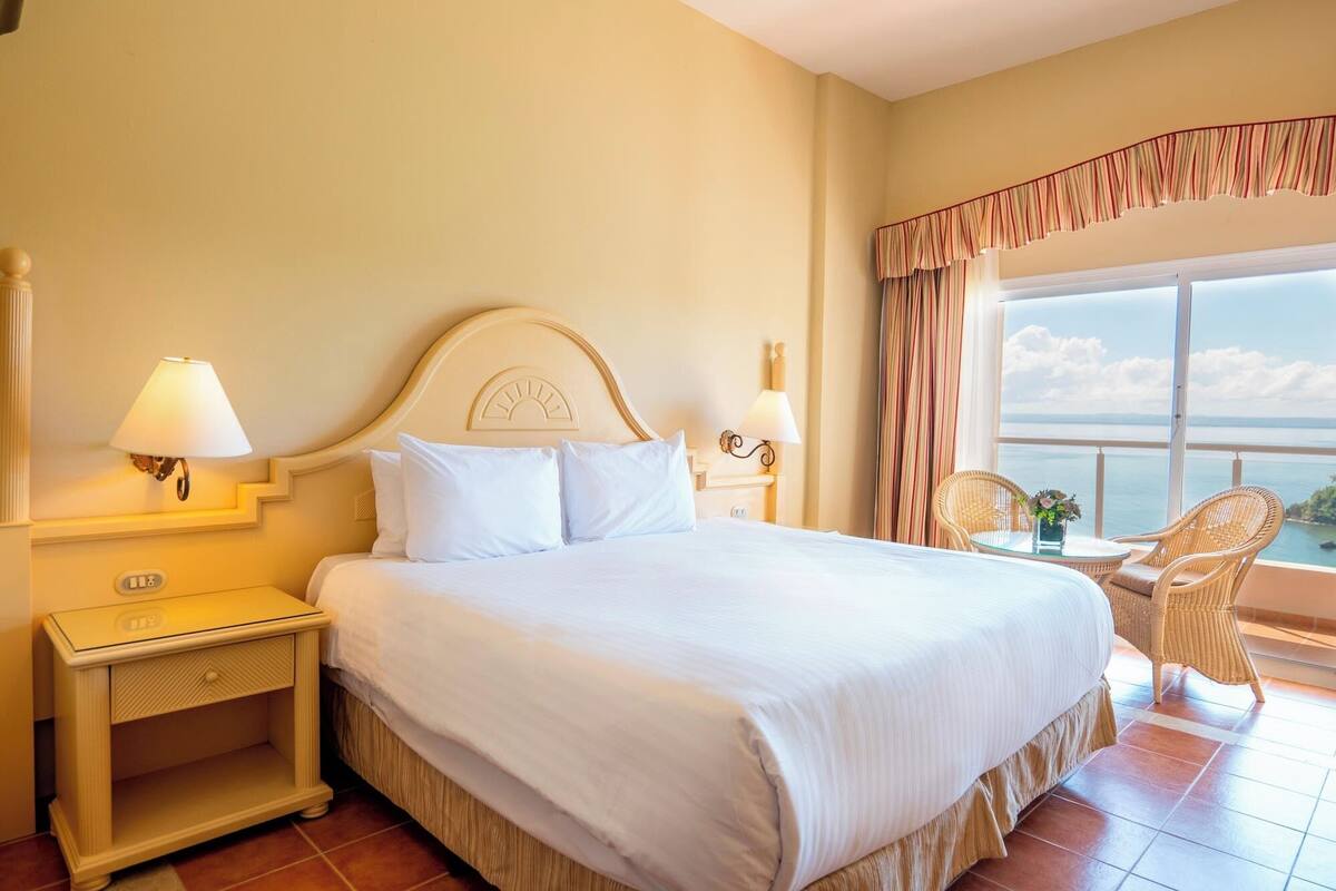 Ein Hotelzimmer mit einem großen Bett, zwei Nachttischen mit Lampen, einem Balkon mit Korbstühlen und Blick aufs Meer.