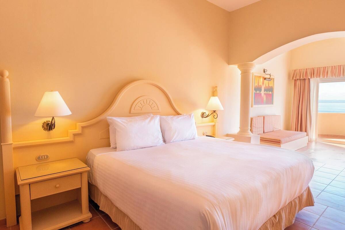 Ein Hotelzimmer mit einem großen Bett, zwei Nachttischen mit Lampen, einer kleinen Sitzecke mit einer Bank und Meerblick vom Balkon.