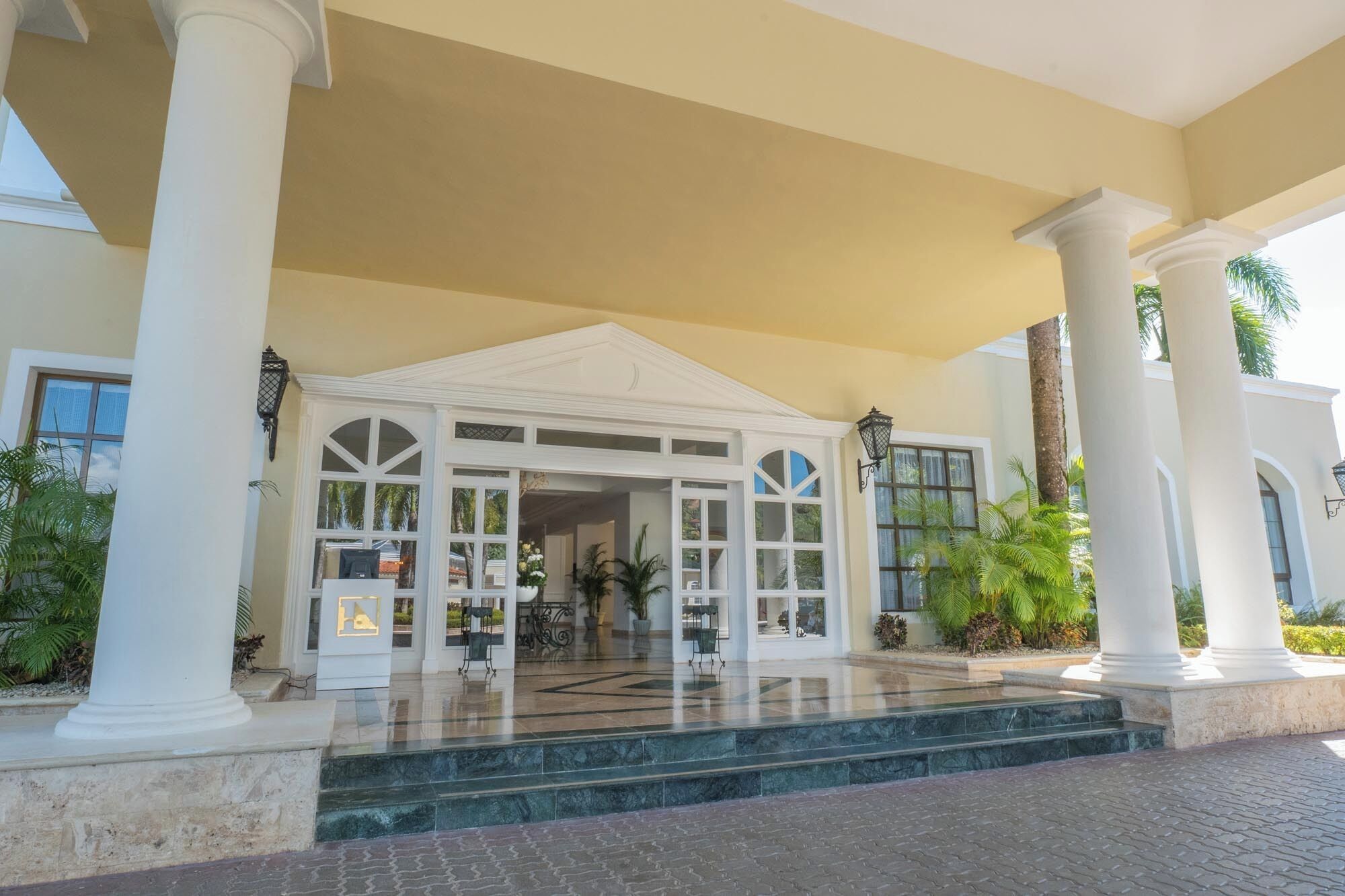 A grand entrance with a covered porch, large columns, and a tiled floor leading to a building with multiple windows and doors.