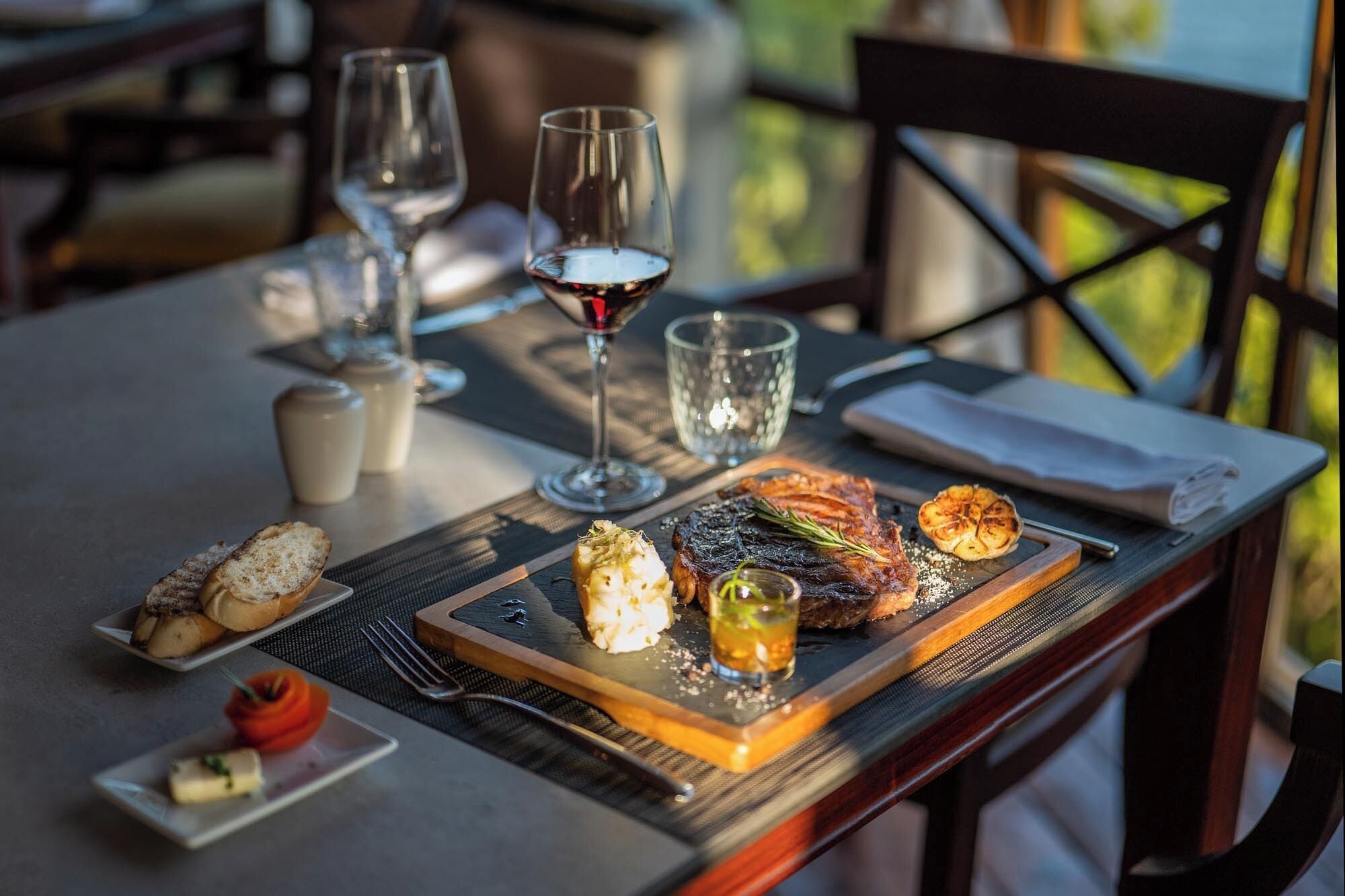 A dining table with a steak, roasted potatoes, and a small dish of sauce. There are also two wine glasses and a glass of water.