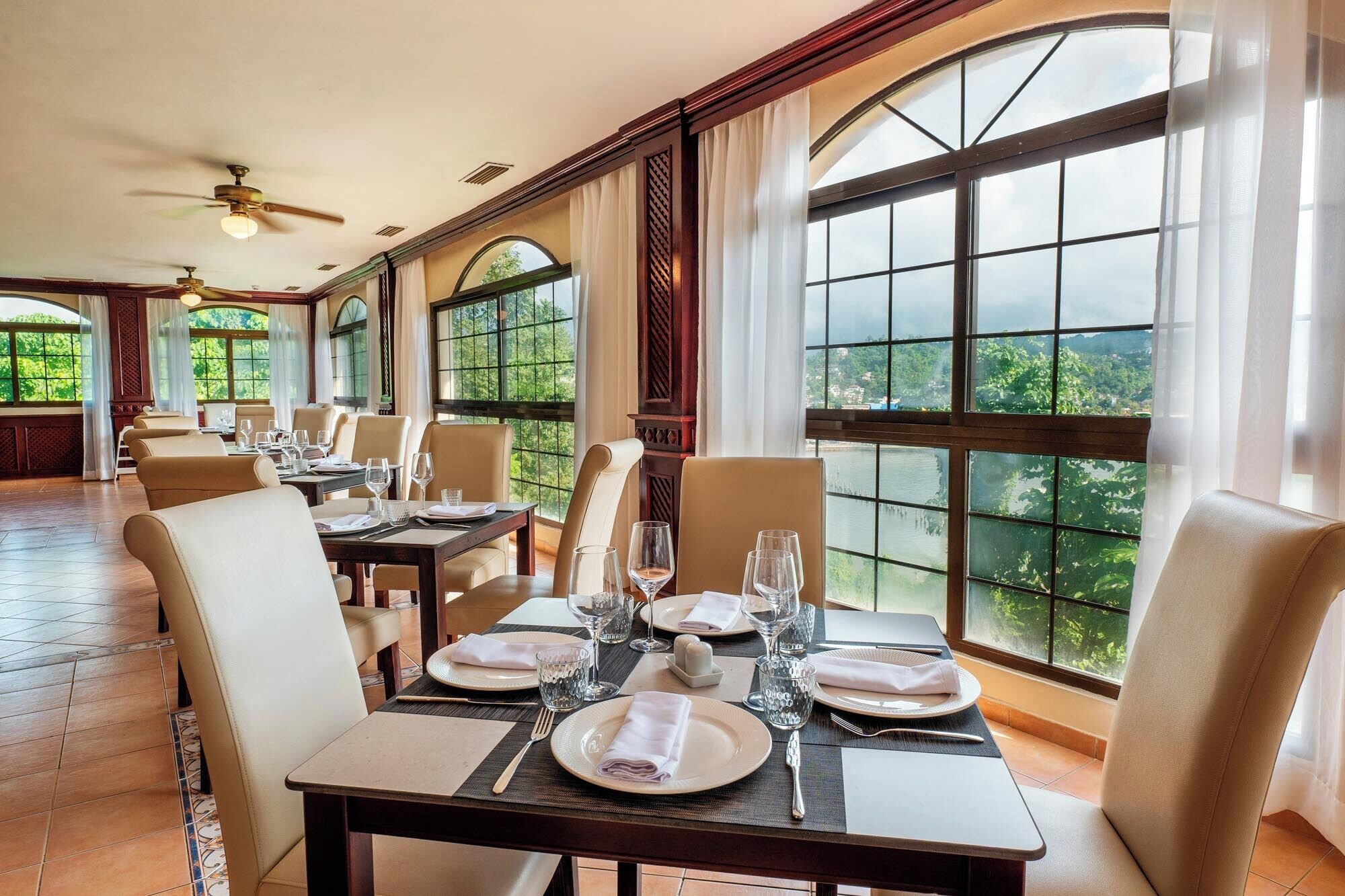 Un comedor con mesas dispuestas para una comida, amplias ventanas que ofrecen vistas a zonas verdes y ventiladores de techo.