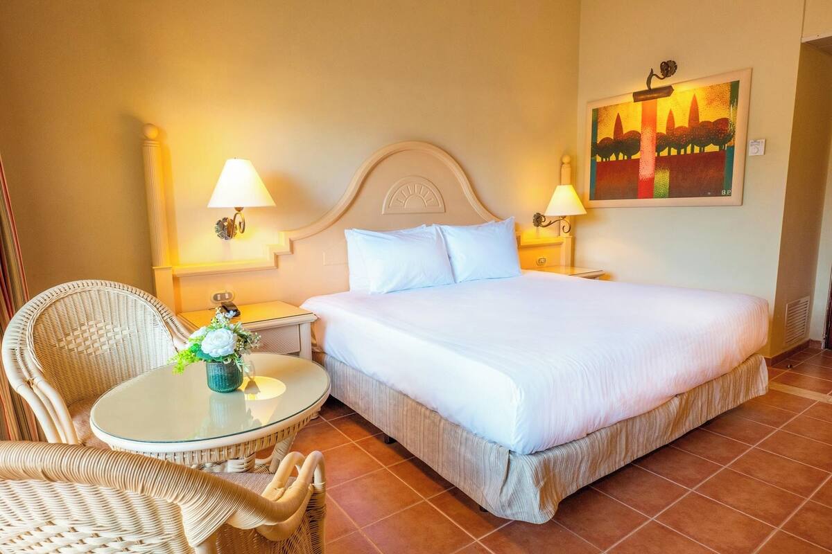 Ein Hotelzimmer mit einem großen Bett, zwei Nachttischlampen, einem Korbstuhl, einem kleinen runden Tisch mit einer Vase und einem Gemälde an der Wand.