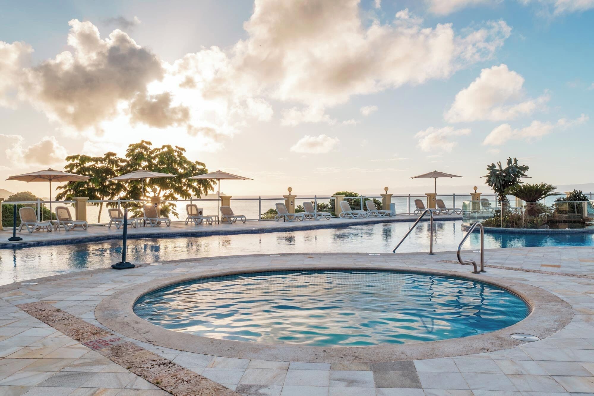 A pool area with lounge chairs and umbrellas, surrounded by a stone deck and a view of the sea.