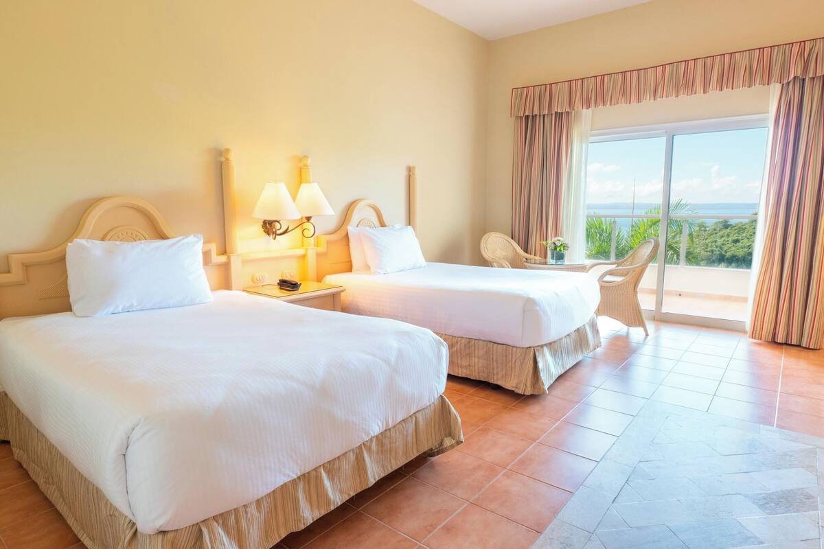 Ein Hotelzimmer mit zwei Betten, einem Balkon mit Vorhängen und Meerblick.