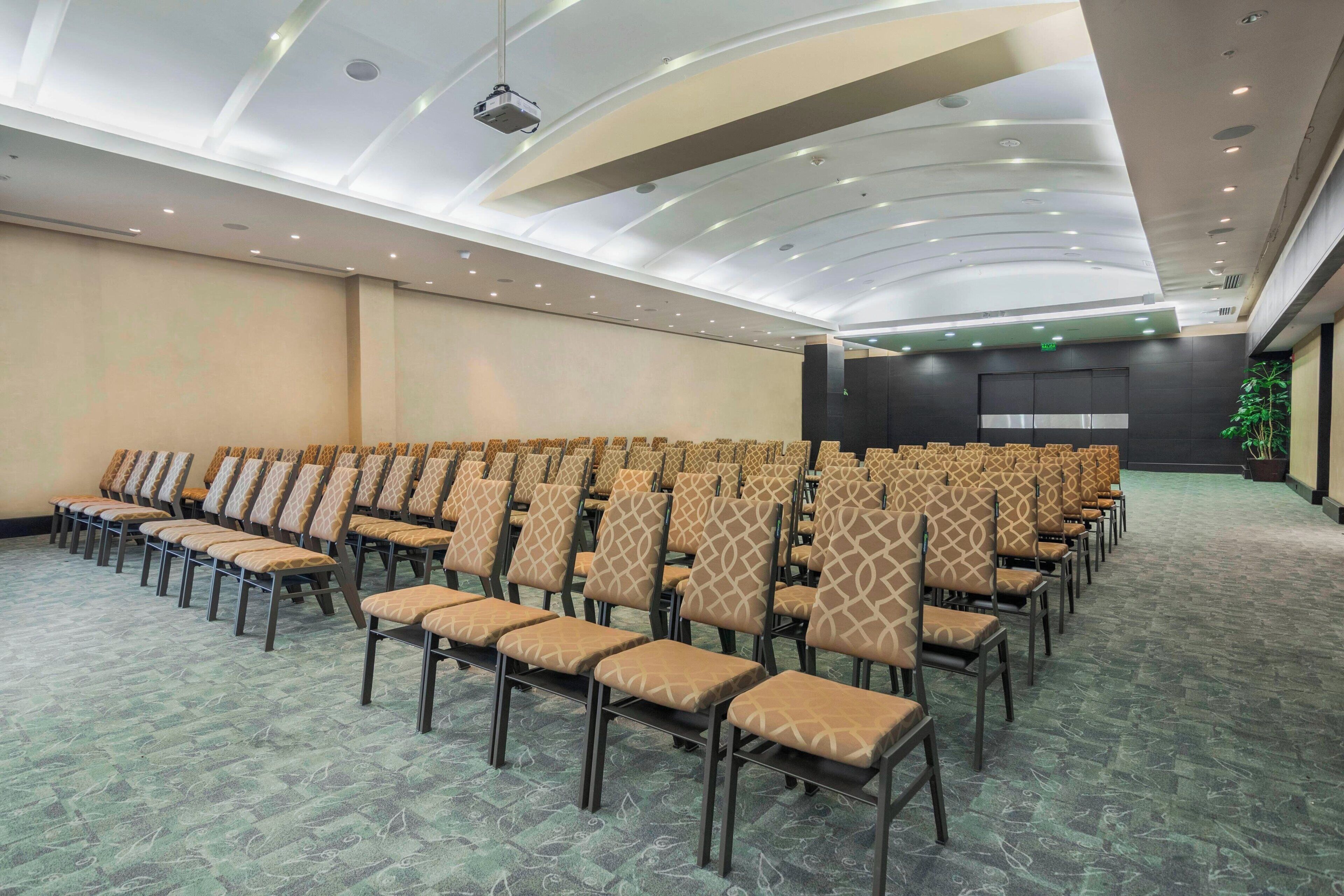 Sala de conferências com fileiras de cadeiras, um projetor e um palco.