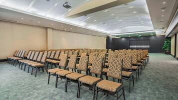 Sala de conferências com fileiras de cadeiras, um projetor e um palco.