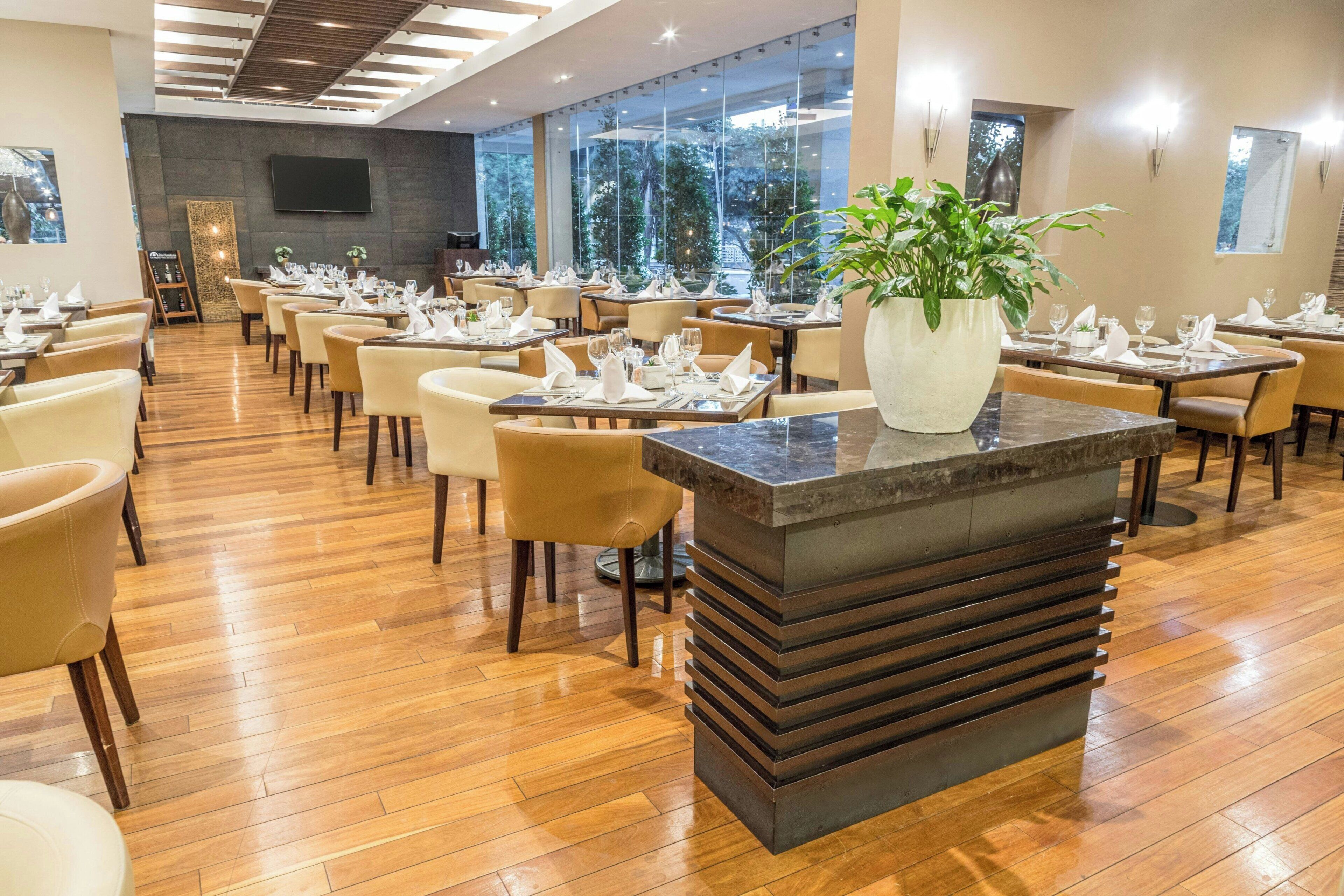 Un'ampia sala ristorante con pavimento in legno, tavoli ordinatamente disposti con tovaglie bianche e una pianta al centro su un bancone in marmo.