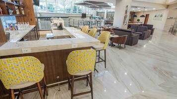 Um amplo hall de hotel com uma receção em mármore, cadeiras com padrão amarelo e uma área de estar moderna com sofás e mesas.
