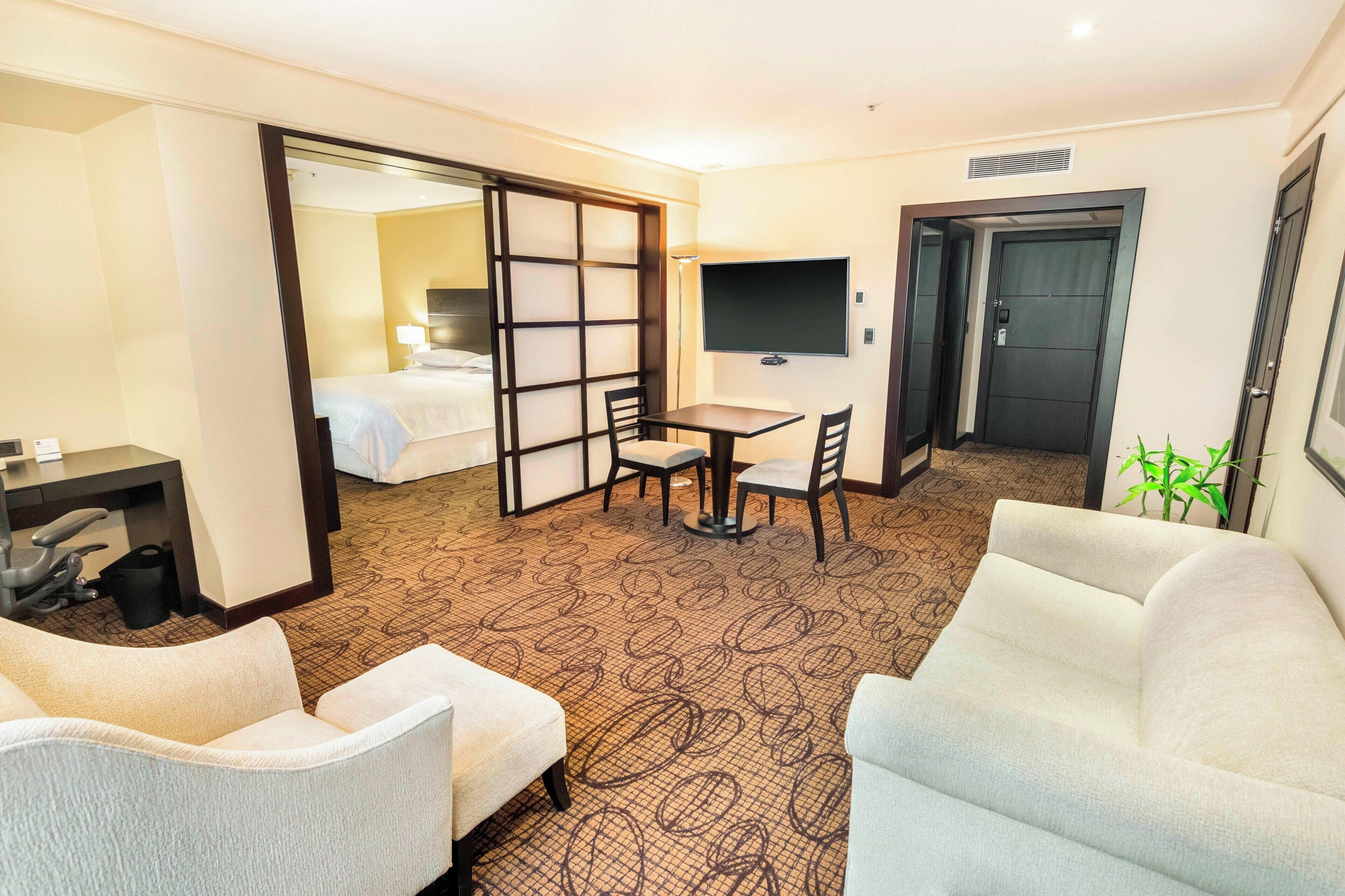 Une chambre d’hôtel comprenant un lit, un bureau, une télévision, un canapé et une porte coulissante donnant sur une autre pièce.