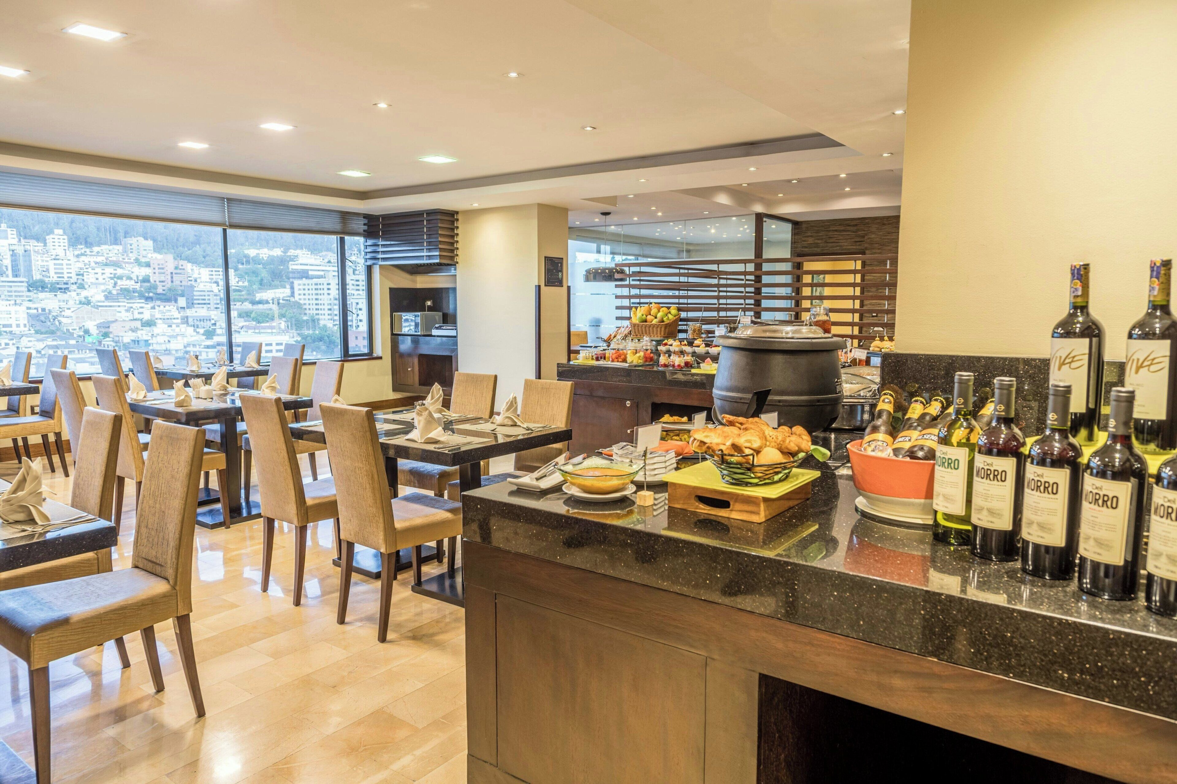 A modern hotel dining area with a buffet setup, a bar with wine bottles, and a view of the cityscape through large windows.