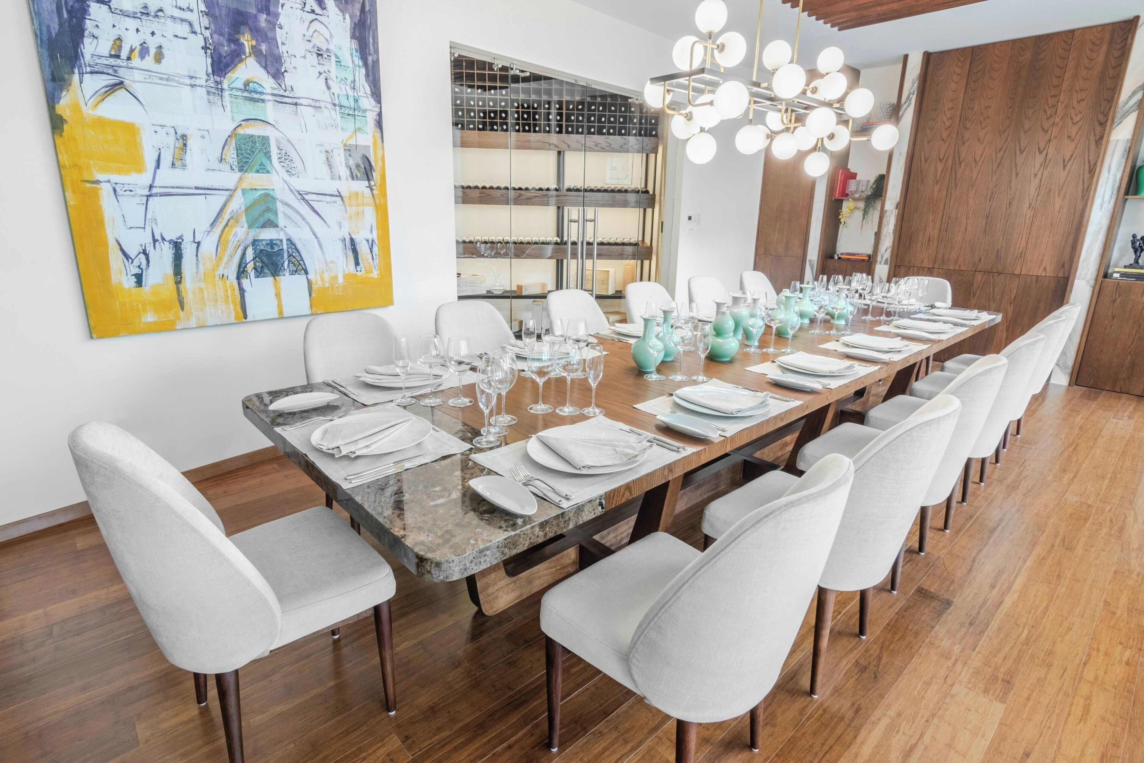 A long dining table set for six with white chairs, a modern chandelier, and a large abstract painting on the wall.