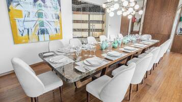 A long dining table set for six with white chairs, a modern chandelier, and a large abstract painting on the wall.