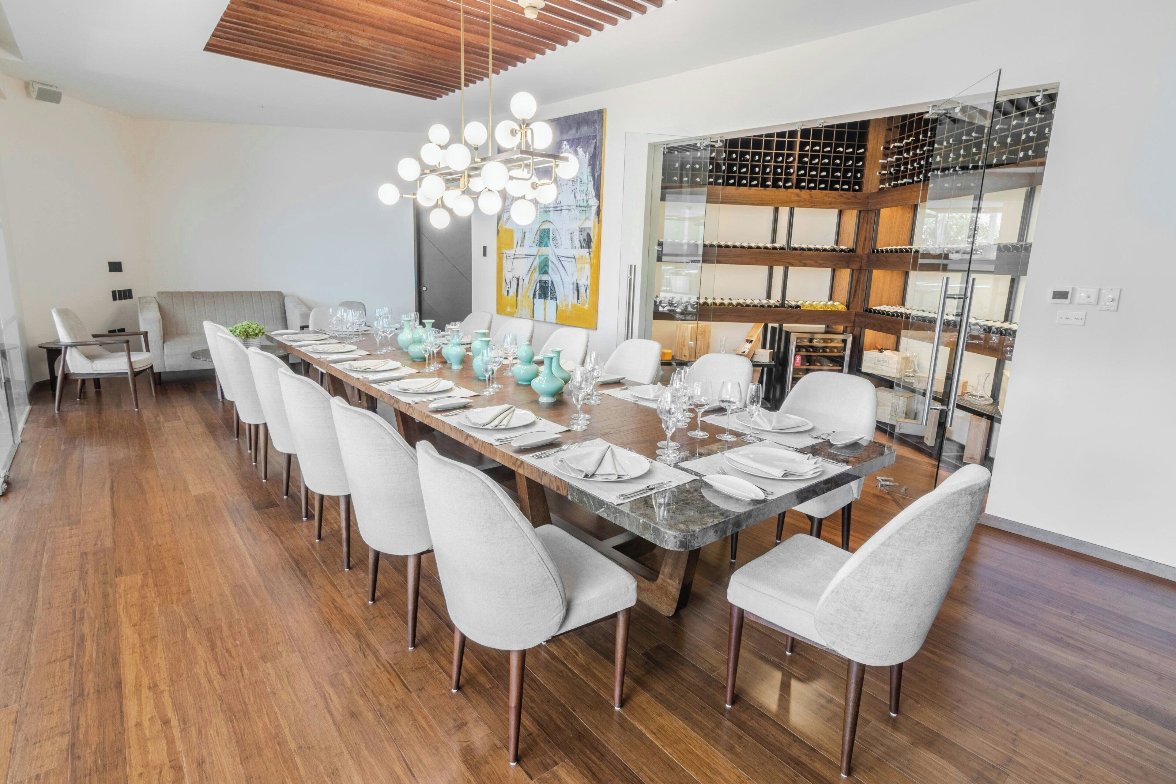 Un espace de salle à manger moderne, avec une longue table dressée pour six personnes, entourée de chaises blanches. Un grand lustre est suspendu au-dessus, et on aperçoit un meuble à vin à l’arrière-plan.