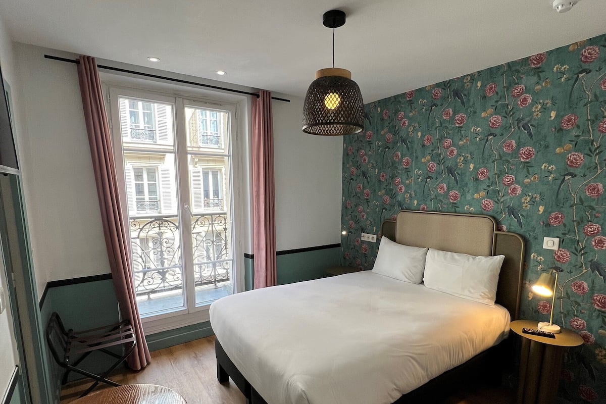 A bedroom with a bed, a small round table, a chair, a nightstand with a lamp, a window with curtains, and a floral wallpaper.