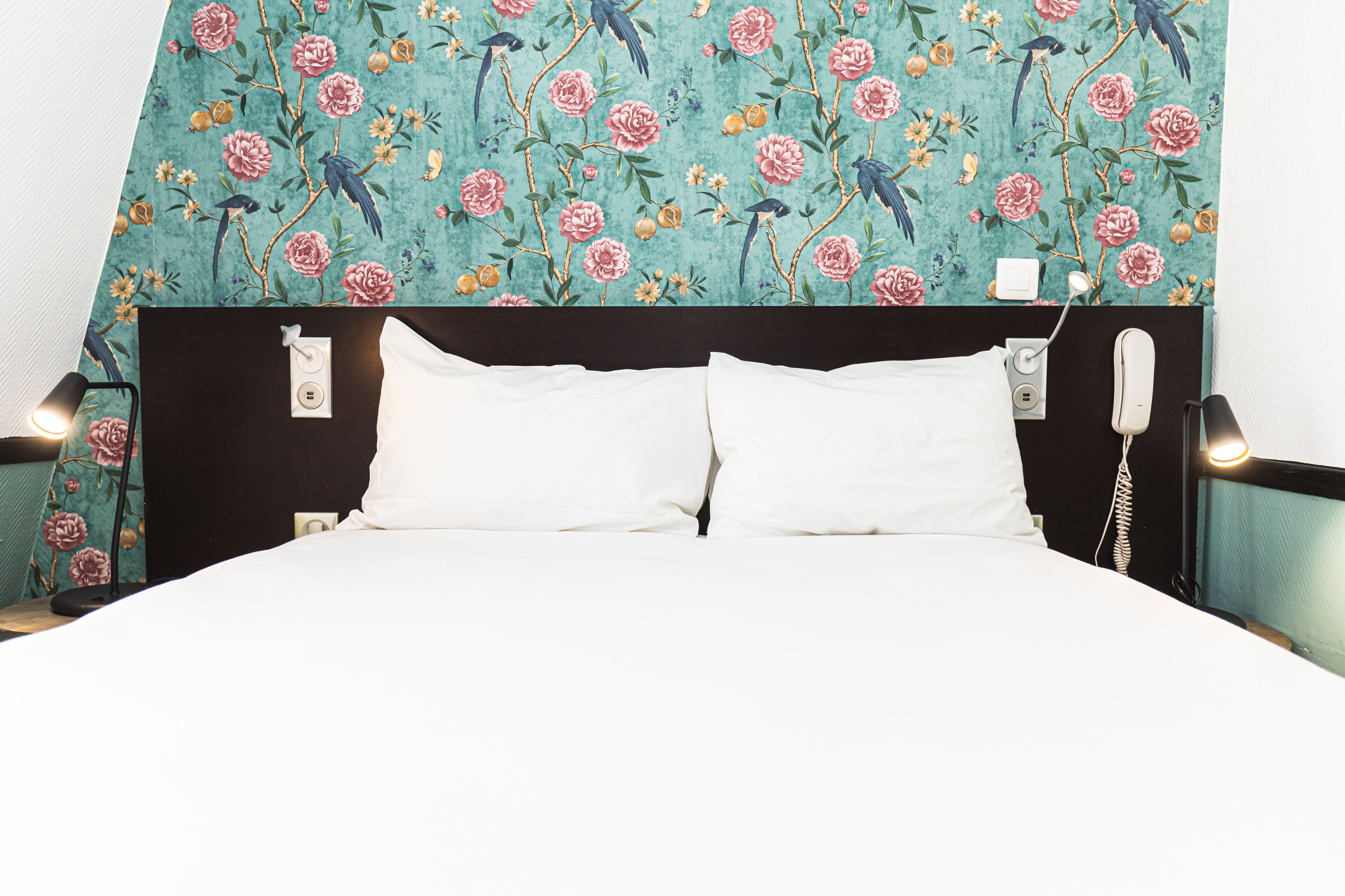 Un lit avec du linge de lit blanc, une tête de lit sombre et un mur orné d’un motif floral.