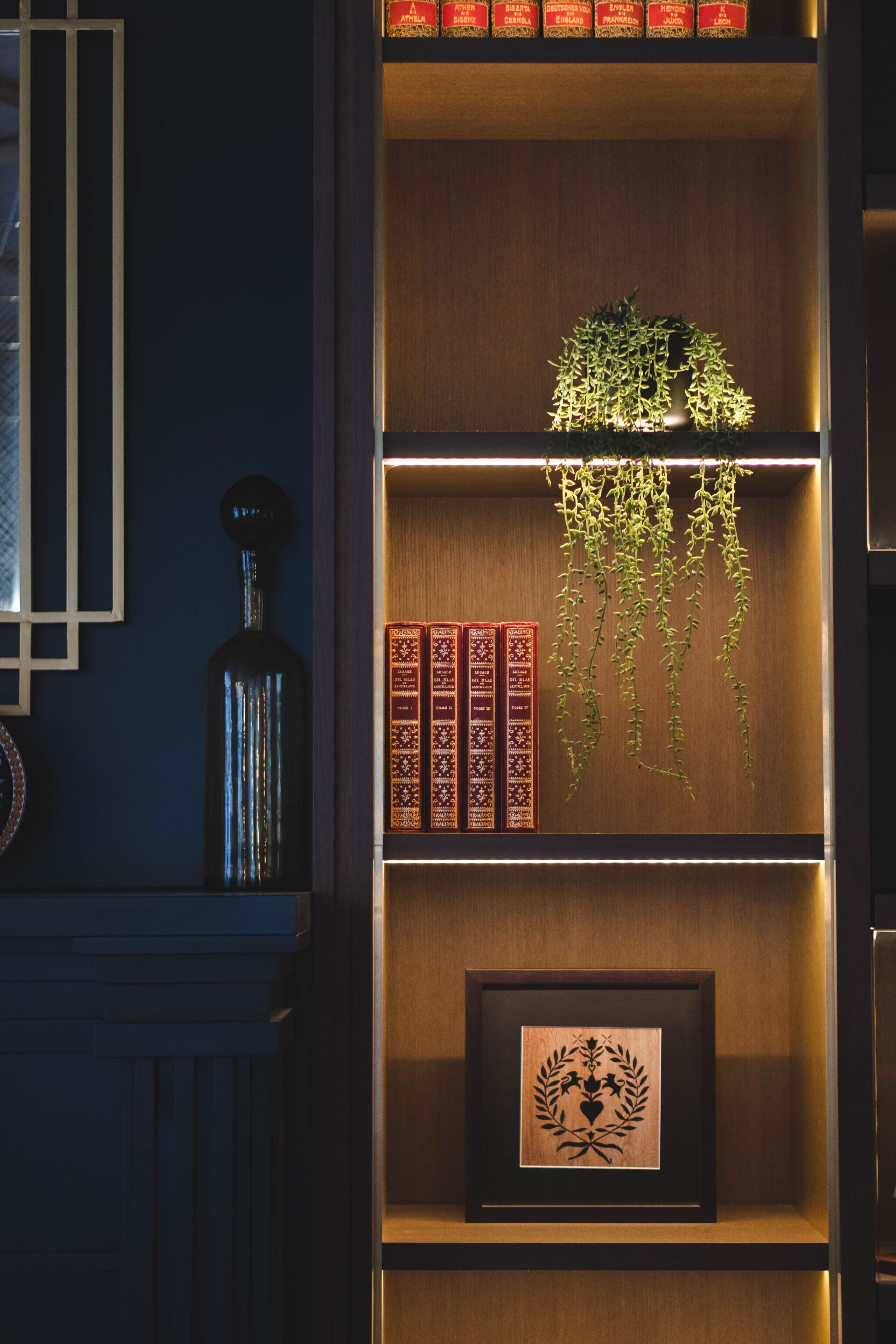 Ein Bücherregal aus Holz mit Büchern, einem gerahmten Kunstwerk und einer hängenden Pflanze.