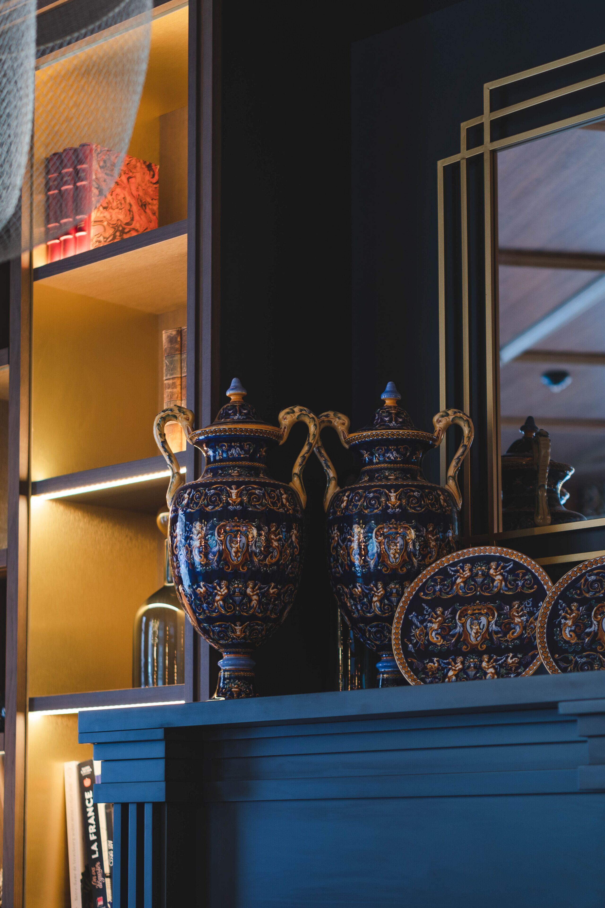 Um quarto escuro com vasos azuis e dourados elaborados em uma estante, um espelho refletindo o quarto e livros na estante.