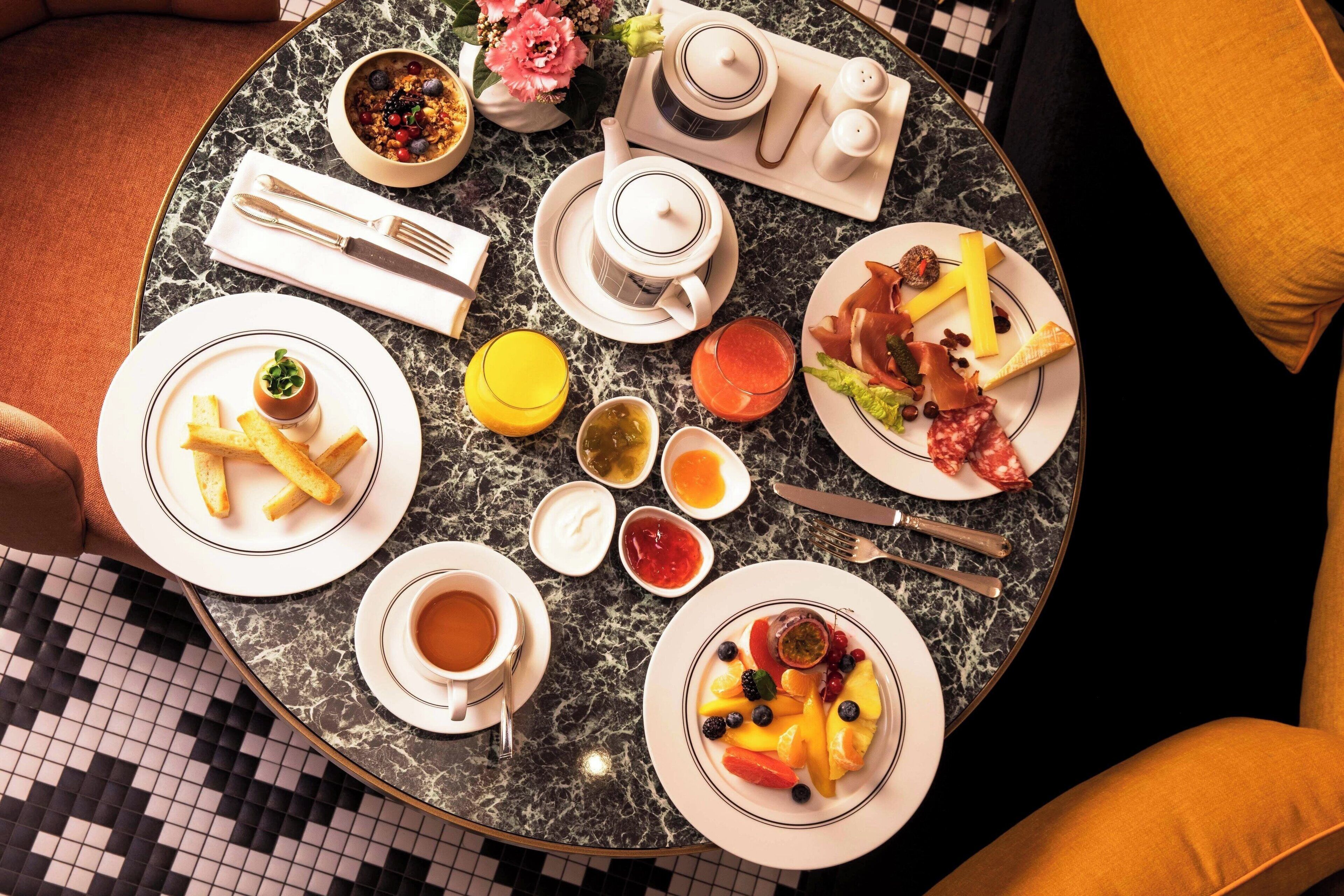A round table with a marble top, surrounded by various breakfast items including plates of food, cups of beverages, and a tea set.