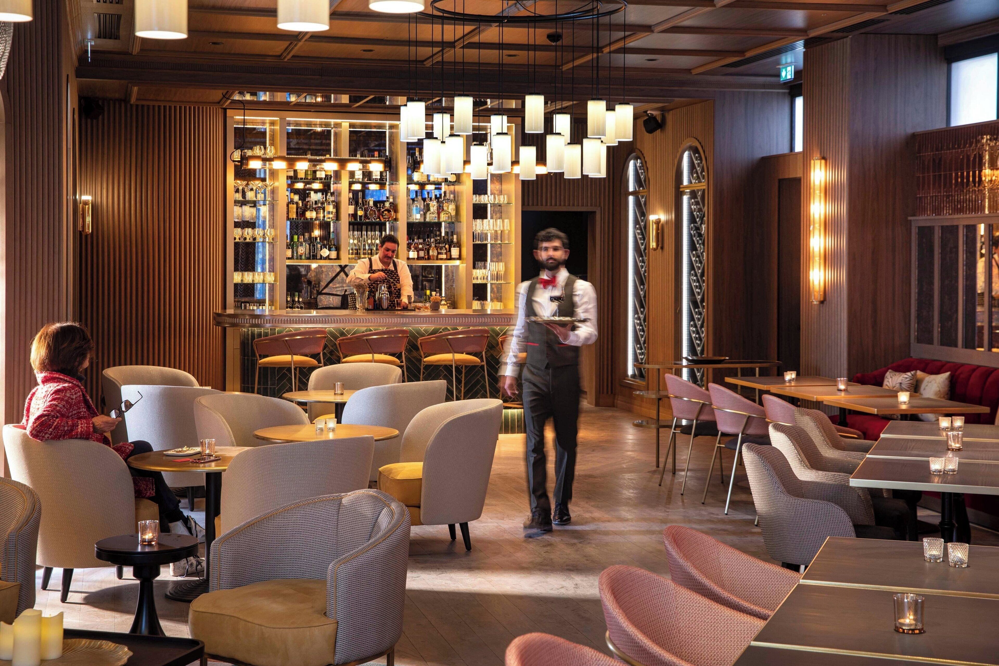 Un restaurant doté d’un bar, de plusieurs types de tables, dont des tables rondes, et de chaises rembourrées, ainsi que d’un serveur qui prend la commande d’un client.