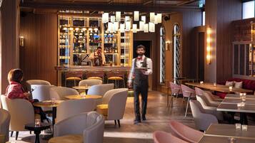 Un restaurante con zona de bar, diversas opciones de asientos, incluyendo mesas redondas y sillas acolchadas, y un camarero atendiendo a un cliente.