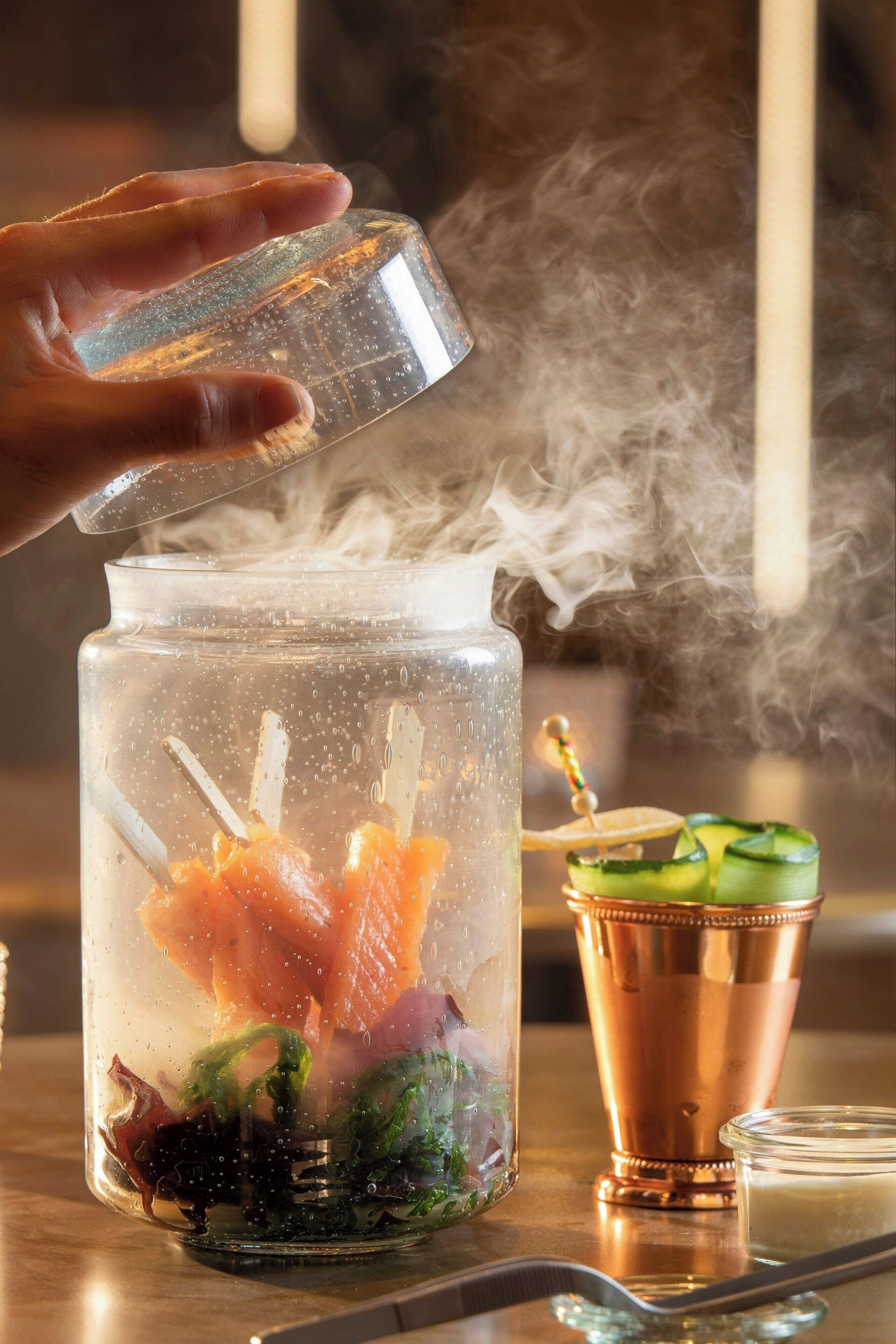 Eine Hand gießt eine klare Flüssigkeit in ein Glasgefäß mit Eis und Lebensmitteln, Dampf steigt auf, und ein Kupfer-Cocktailshaker sowie ein Glas befinden sich auf dem Tisch.