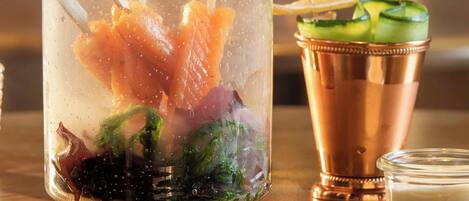 A hand pouring a clear liquid into a glass jar with ice and food items, steam rising, and a copper cocktail shaker and glass on the table.