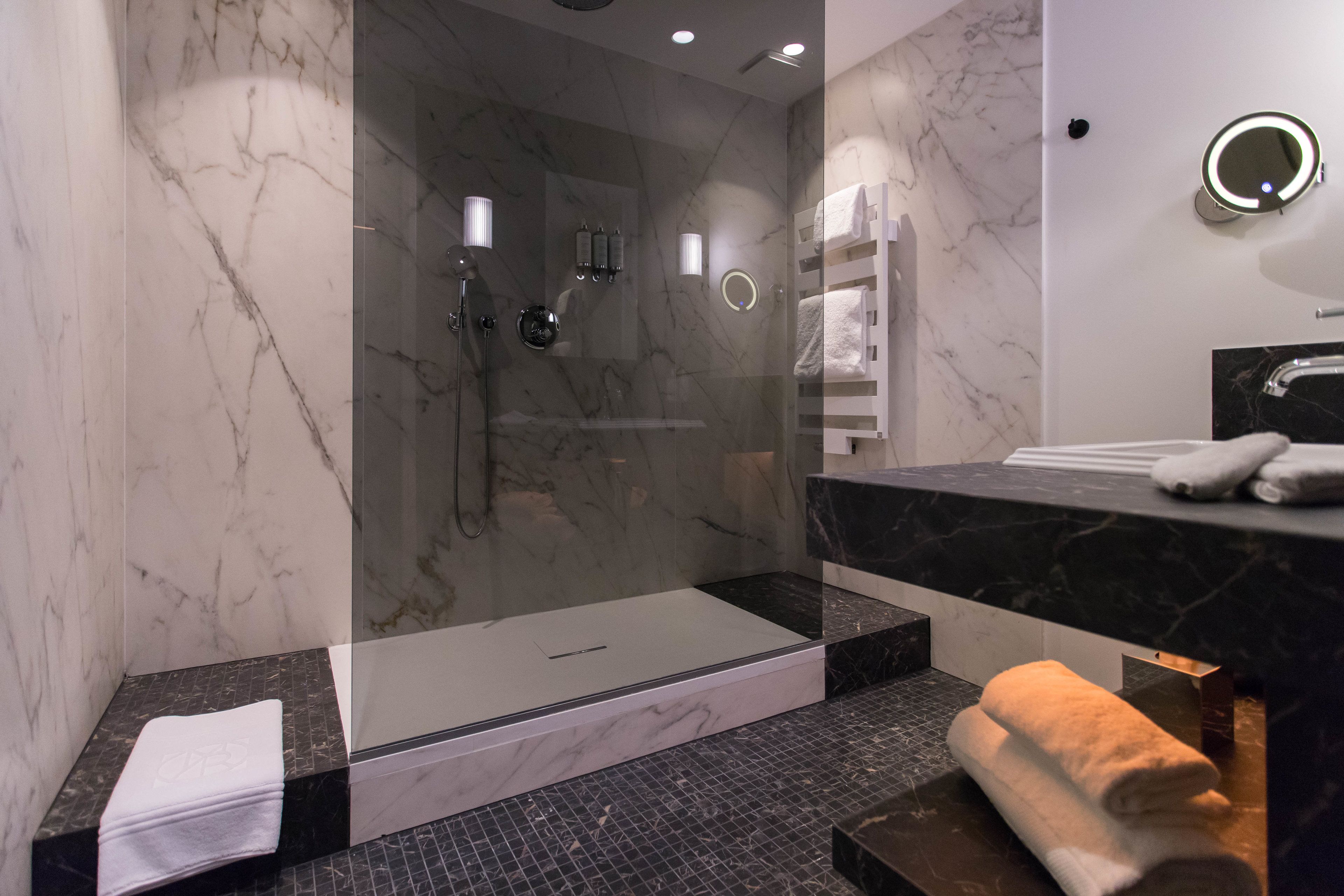 Un baño moderno con ducha envuelta en cristal, paredes de mármol y encimera negra con toallero.