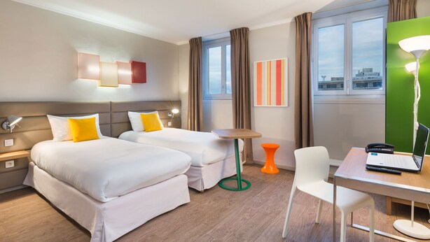 A hotel room with two beds, a desk with a laptop, a chair, a small table, and a lamp.